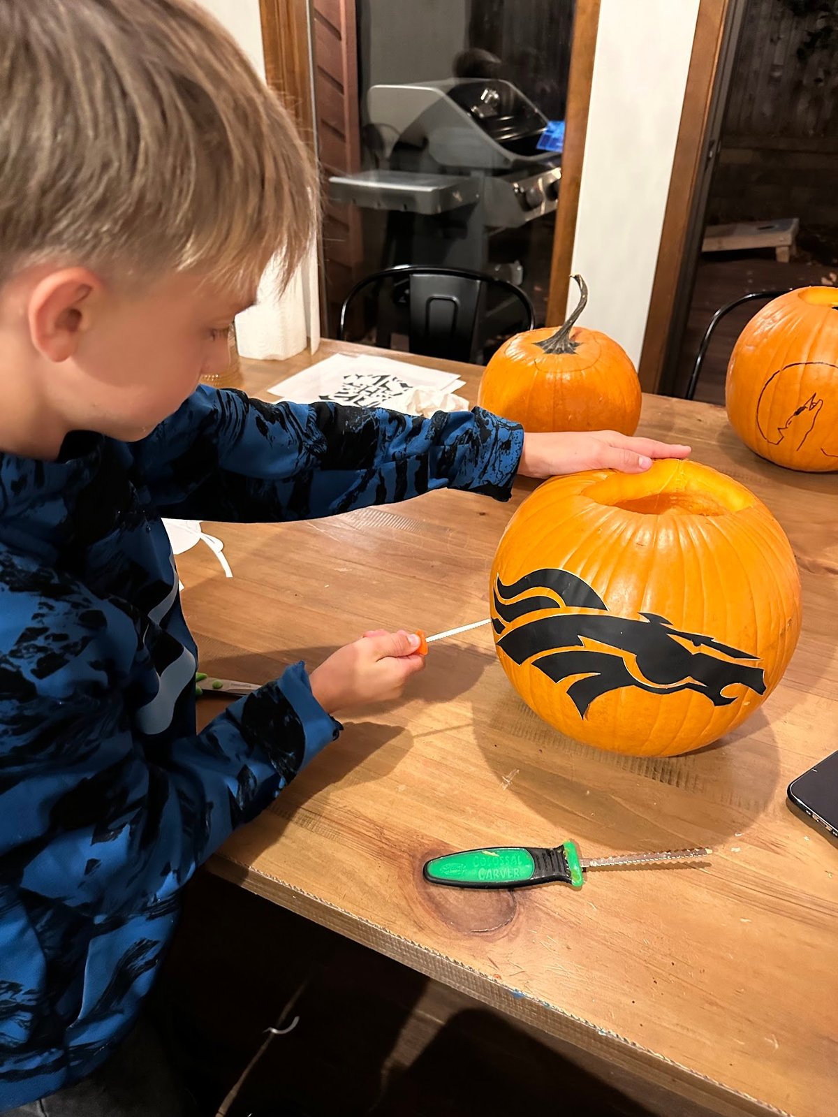 Child in blue jacket painting a black lightning bolt design on an orange pumpkin at a wooden table